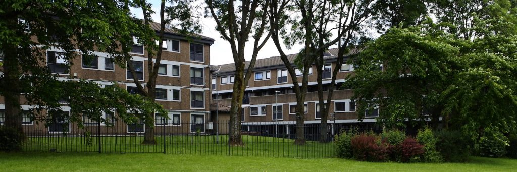 Photo of two low-rise blocks of flats, Quinton and Pershore, with open green space and trees in the foreground. The blocks are part of the Lower Falinge estate in Rochdale.